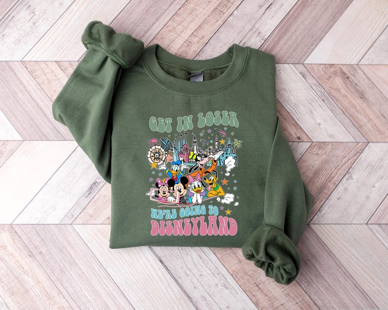May include: Olive green sweatshirt with the text "GET IN LOSER WE'RE GOING TO DISNEYLAND" in a retro font. The graphic features cartoon characters and a colorful illustration of a theme park.