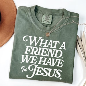 K&ouml;nnte beinhalten: Ein gr&uuml;nes T-Shirt mit dem Text "What a Friend We Have in Jesus" in wei&szlig;en Buchstaben. Das Shirt hat einen Used-Look und ist aus einem weichen, bequemen Stoff.