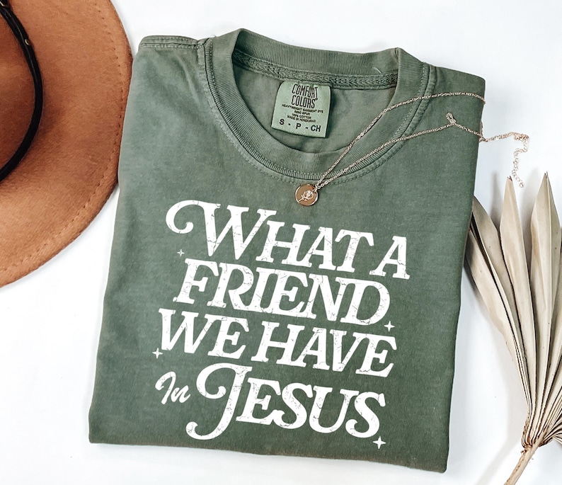 K&ouml;nnte beinhalten: Ein gr&uuml;nes T-Shirt mit dem Text "What a Friend We Have in Jesus" in wei&szlig;en Buchstaben. Das Shirt hat einen Used-Look und ist aus einem weichen, bequemen Stoff.
