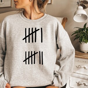May include: A light gray sweatshirt with a black tally mark design. The design consists of vertical lines with diagonal lines through them, representing a count. The sweatshirt is a casual, comfortable style.