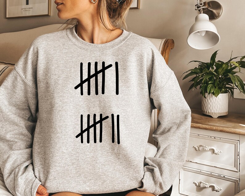 May include: A light gray sweatshirt with a black tally mark design. The design consists of vertical lines with diagonal lines through them, representing a count. The sweatshirt is a casual, comfortable style.