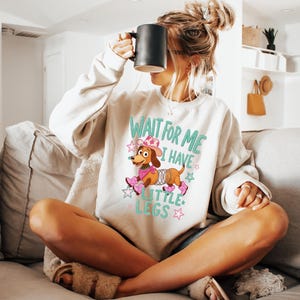 Puede incluir: Una sudadera beige de cuello redondo con un gr&aacute;fico de un perro salchicha marr&oacute;n que lleva un sombrero de vaquero rosa, un pa&ntilde;uelo rosa y botas rosas. El perro tiene un cuerpo de resorte plateado. El texto dice WAIT FOR ME I HAVE LITTLE LEGS en turquesa.