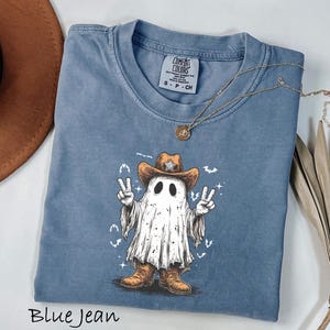 Pu&ograve; includere: T-shirt blu jeans con un fantasma dei cartoni animati con cappello e stivali da cowboy, che fa il segno della pace. La maglietta ha un girocollo e maniche corte. Una collana d'oro con un ciondolo &egrave; drappeggiata sulla maglietta. La scritta "Blue Jean" &egrave; in basso.