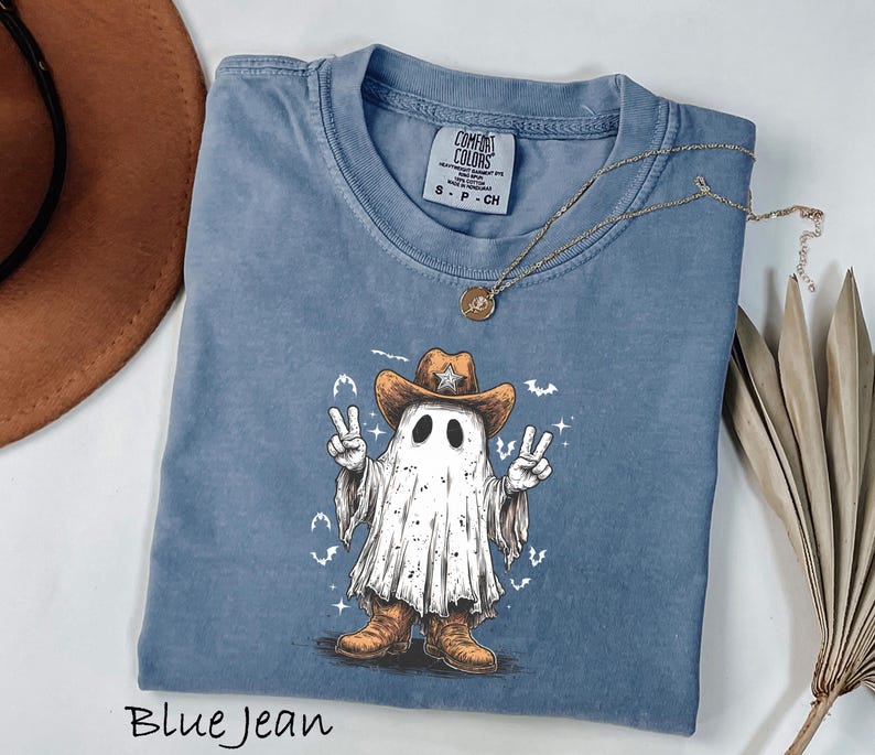 Pu&ograve; includere: T-shirt blu jeans con un fantasma dei cartoni animati con cappello e stivali da cowboy, che fa il segno della pace. La maglietta ha un girocollo e maniche corte. Una collana d'oro con un ciondolo &egrave; drappeggiata sulla maglietta. La scritta "Blue Jean" &egrave; in basso.