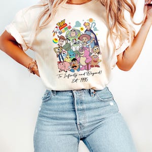 May include: Cream-colored t-shirt featuring a colorful graphic of Toy Story characters. The design includes Woody, Buzz Lightyear, and others, with the text "Toy Story Land" and "To Infinity and Beyond! Est. 1995".