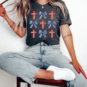 May include: A charcoal grey t-shirt featuring a pattern of red crosses and blue bows. The shirt is being worn by a person with long blonde hair.