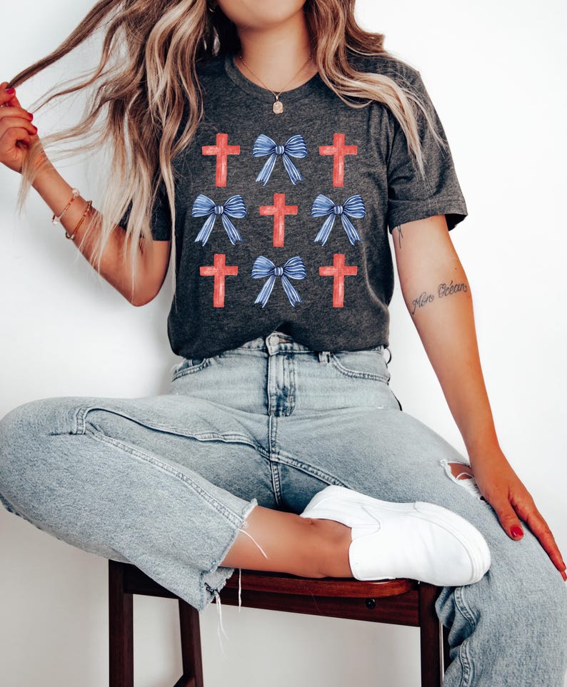 May include: A charcoal grey t-shirt featuring a pattern of red crosses and blue bows. The shirt is being worn by a person with long blonde hair.