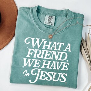 K&ouml;nnte beinhalten: Ein blaugr&uuml;nes T-Shirt mit einem wei&szlig;en, abgenutzten Aufdruck, der "What a Friend We Have in Jesus" lautet.