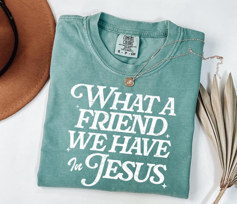 K&ouml;nnte beinhalten: Ein blaugr&uuml;nes T-Shirt mit einem wei&szlig;en, abgenutzten Aufdruck, der "What a Friend We Have in Jesus" lautet.