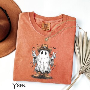 Pu&ograve; includere: Una maglietta arancione bruciato con una grafica di un fantasma con cappello e stivali da cowboy, che fa il segno della pace. La maglietta ha un girocollo e maniche corte. Una collana d'oro e un cappello da cowboy marrone sono anche nell'immagine.