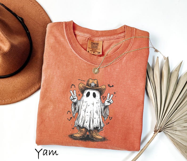 Pu&ograve; includere: Una maglietta arancione bruciato con una grafica di un fantasma con cappello e stivali da cowboy, che fa il segno della pace. La maglietta ha un girocollo e maniche corte. Una collana d'oro e un cappello da cowboy marrone sono anche nell'immagine.