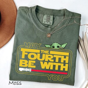 May include: Moss green t-shirt with the text "MAY THE FOURTH BE WITH YOU" in yellow and white, featuring a cartoon character. The design includes a red lightsaber and a yellow border. A gold necklace is draped across the shirt.