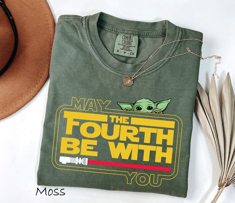 May include: Moss green t-shirt with the text "MAY THE FOURTH BE WITH YOU" in yellow and white, featuring a cartoon character. The design includes a red lightsaber and a yellow border. A gold necklace is draped across the shirt.
