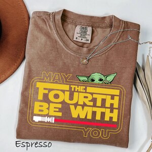 May include: Espresso-colored t-shirt with the text "MAY THE FOURTH BE WITH YOU" in yellow and white, featuring a green character graphic. The shirt has a crew neck and a small tag. A silver necklace with a pendant is also visible.