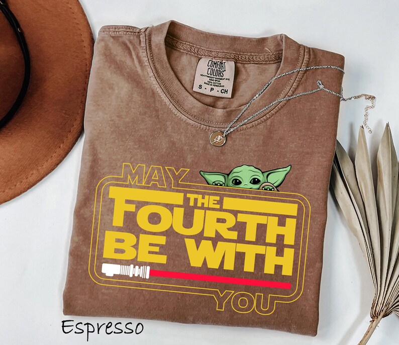 May include: Espresso-colored t-shirt with the text "MAY THE FOURTH BE WITH YOU" in yellow and white, featuring a green character graphic. The shirt has a crew neck and a small tag. A silver necklace with a pendant is also visible.