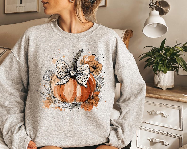 May include: A light gray sweatshirt featuring a watercolor-style pumpkin design. The pumpkin is orange with a black and white polka dot bow and surrounded by floral accents in shades of orange and white. A fall-themed apparel item.