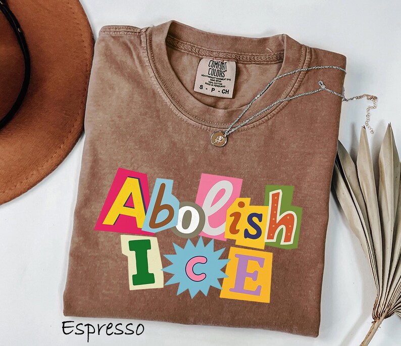May include: Espresso-colored t-shirt with the words "Abolish ICE" in a colorful, cut-out style. The shirt has a crew neck and a small tag on the collar. A silver necklace with a pendant is draped across the shirt. A brown hat and dried plant are in the background.
