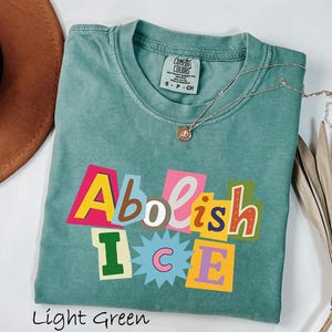 May include: Light green t-shirt with the words "Abolish ICE" in a colorful, cut-out style design. The shirt has a crew neck and short sleeves. A gold necklace with a small pendant is draped across the shirt. The shirt is folded on a white surface.