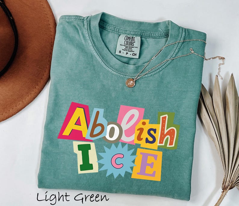 May include: Light green t-shirt with the words "Abolish ICE" in a colorful, cut-out style design. The shirt has a crew neck and short sleeves. A gold necklace with a small pendant is draped across the shirt. The shirt is folded on a white surface.