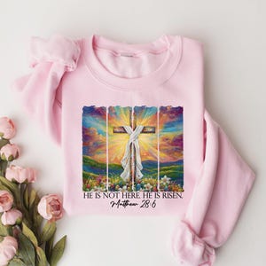 May include: Pink sweatshirt featuring a colourful painting of a cross with the text "He is not here. He is risen. Matthew 28:6"