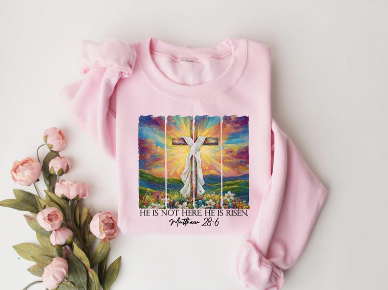 May include: Pink sweatshirt featuring a colourful painting of a cross with the text "He is not here. He is risen. Matthew 28:6"