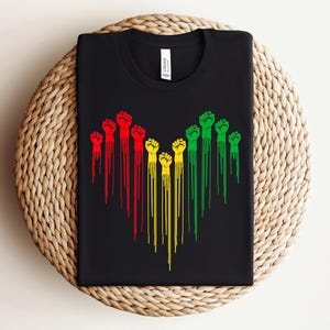 May include: Black t-shirt featuring a graphic of stylized fists in red, yellow, and green, with dripping lines extending downward. The design is centered on the shirt, creating a bold visual statement. The shirt is folded neatly.