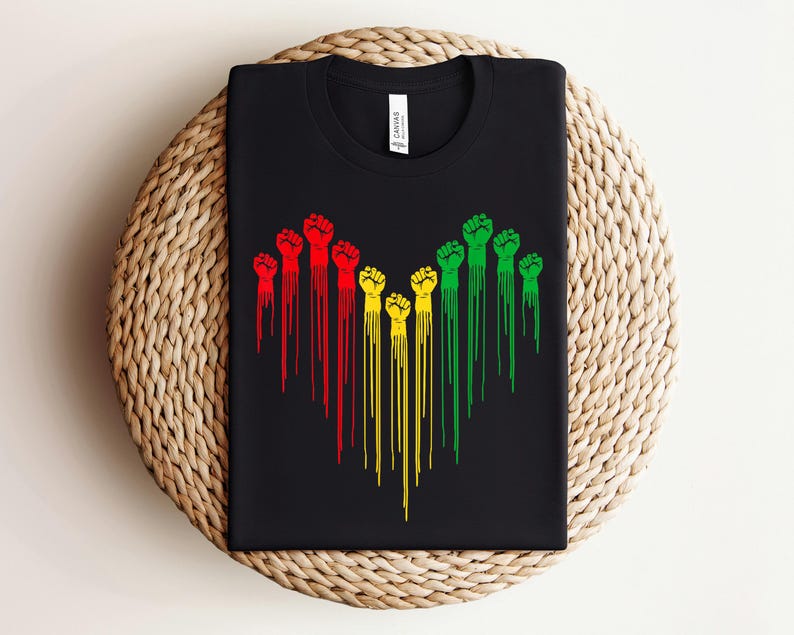 May include: Black t-shirt featuring a graphic of stylized fists in red, yellow, and green, with dripping lines extending downward. The design is centered on the shirt, creating a bold visual statement. The shirt is folded neatly.