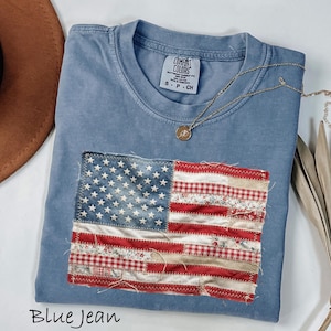 May include: Blue jean colored t-shirt with a sewn-on American flag design. The flag is made of various fabrics, including denim and patterned red and white fabrics. A gold necklace with a pendant rests on the shirt. The shirt has a Comfort Colors tag.