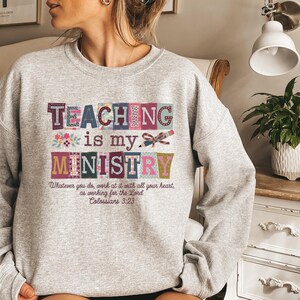 May include: A light gray sweatshirt with the words "TEACHING is my MINISTRY" in colorful block letters. Below the text is the Bible verse Colossians 3:23. The sweatshirt is a comfortable, casual style.