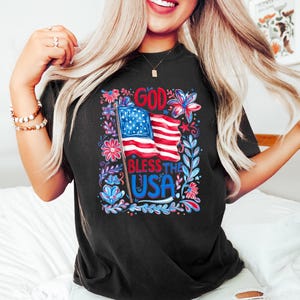 May include: A black short-sleeved t-shirt featuring a graphic print of an American flag on a pole, surrounded by red, white, and blue floral illustrations. The text "GOD BLESS THE USA" is printed in bold blue and red lettering on the front.