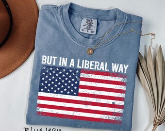 May include: Blue jean colored t-shirt with the text "BUT IN A LIBERAL WAY" above a distressed American flag graphic. A gold necklace with a pendant rests on the shirt. The shirt is a Comfort Colors brand.