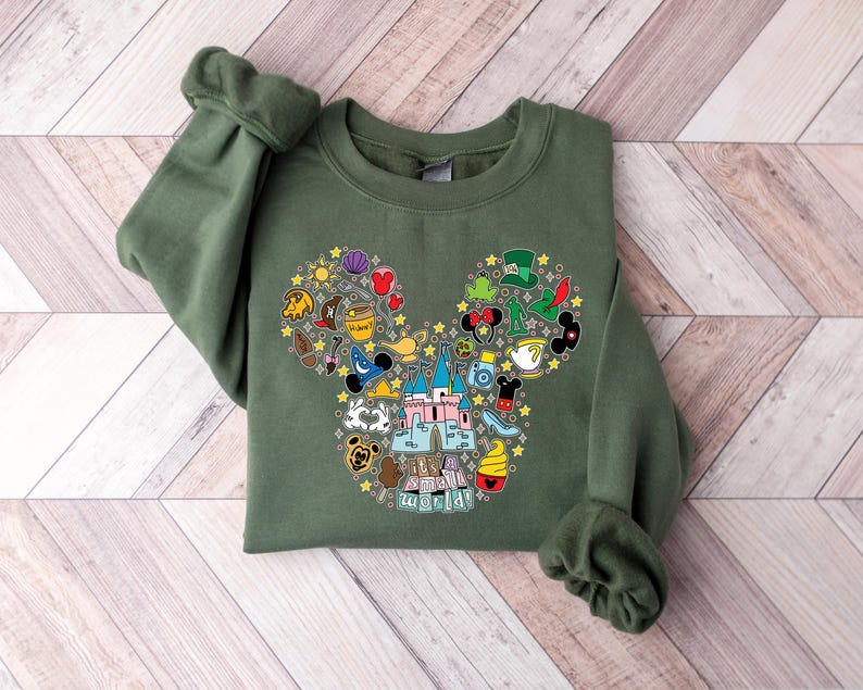 Puede incluir: Una sudadera verde oliva con un dise&ntilde;o de cabeza de Mickey Mouse lleno de coloridos iconos tem&aacute;ticos de Disney. El dise&ntilde;o incluye un castillo, sombreros y varias im&aacute;genes relacionadas con los personajes. La sudadera tiene mangas largas y cuello redondo.