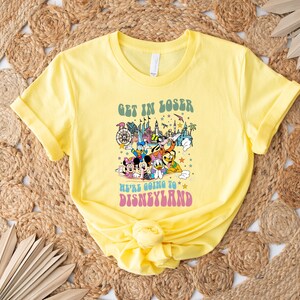 May include: A yellow t-shirt with the text "GET IN LOSER WE'RE GOING TO DISNEYLAND" in green and pink. The shirt features a colorful graphic of Disney characters and landmarks.