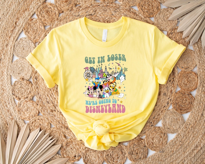 May include: A yellow t-shirt with the text "GET IN LOSER WE'RE GOING TO DISNEYLAND" in green and pink. The shirt features a colorful graphic of Disney characters and landmarks.