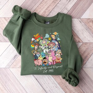 May include: Green sweatshirt featuring a colorful graphic of Toy Story characters. The design includes Buzz Lightyear, Woody, and other characters from the movie. The text "Toy Land" and "To Infinity and Beyond! Est 1995" are also visible.