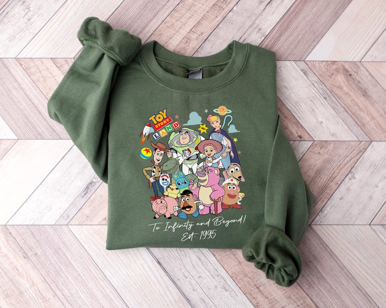 May include: Green sweatshirt featuring a colorful graphic of Toy Story characters. The design includes Buzz Lightyear, Woody, and other characters from the movie. The text "Toy Land" and "To Infinity and Beyond! Est 1995" are also visible.