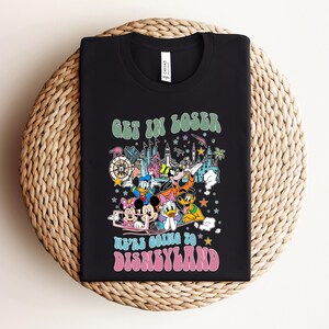 May include: Black t-shirt featuring a colorful graphic of Disney characters and park icons. The text "GET IN LOSER" is above the graphic, and "WE'RE GOING TO DISNEYLAND" is below. The shirt is displayed on a woven surface.