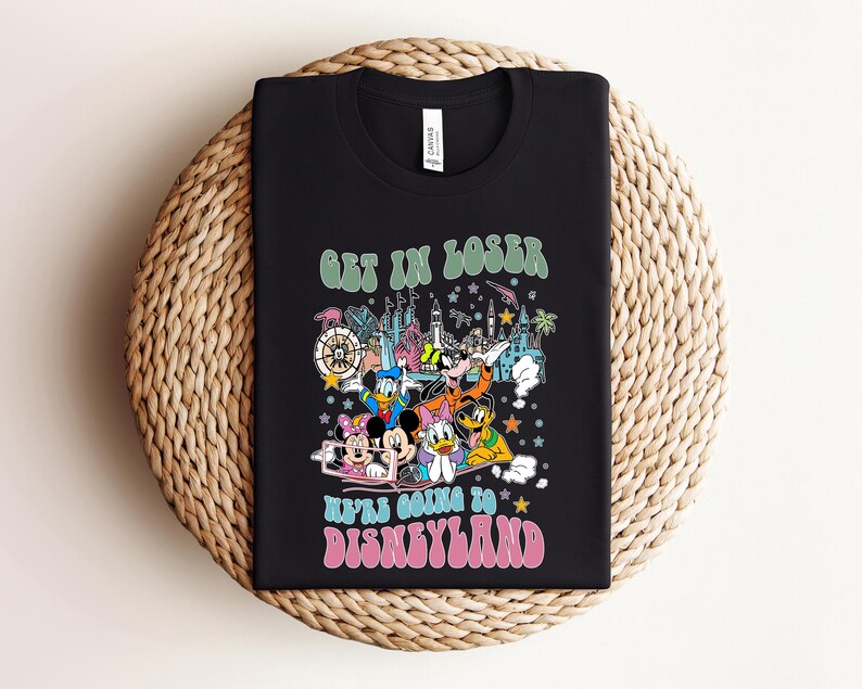 May include: Black t-shirt featuring a colorful graphic of Disney characters and park icons. The text "GET IN LOSER" is above the graphic, and "WE'RE GOING TO DISNEYLAND" is below. The shirt is displayed on a woven surface.