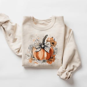 May include: Cream-colored sweatshirt featuring a watercolor-style pumpkin design. The pumpkin is orange with a black and white polka dot bow. Floral accents in orange and gray surround the pumpkin, creating a fall-themed aesthetic.