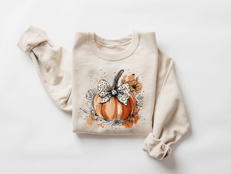 May include: Cream-colored sweatshirt featuring a watercolor-style pumpkin design. The pumpkin is orange with a black and white polka dot bow. Floral accents in orange and gray surround the pumpkin, creating a fall-themed aesthetic.