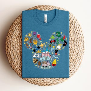 Puede incluir: Camiseta azul con un gr&aacute;fico colorido de orejas de Mickey Mouse llenas de iconos tem&aacute;ticos, incluyendo un castillo, tazas de t&eacute; y estrellas. El texto "It's a small world!" est&aacute; en la parte inferior. La camiseta est&aacute; doblada sobre una superficie tejida.