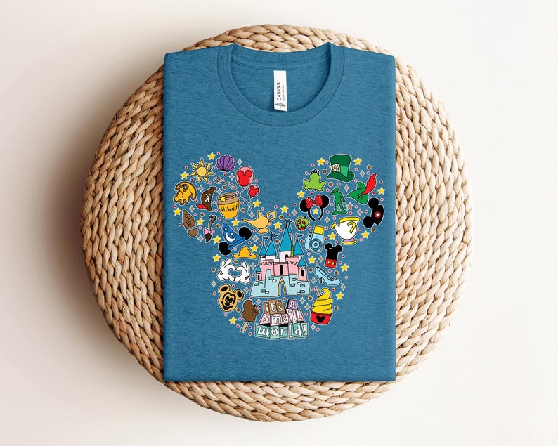 Puede incluir: Camiseta azul con un gr&aacute;fico colorido de orejas de Mickey Mouse llenas de iconos tem&aacute;ticos, incluyendo un castillo, tazas de t&eacute; y estrellas. El texto "It's a small world!" est&aacute; en la parte inferior. La camiseta est&aacute; doblada sobre una superficie tejida.