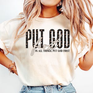 May include: Cream-colored t-shirt with the text "PUT GOD FIRST" in black block letters with star accents. The word "First" is in a cursive font. Below the text is the phrase "IN ALL THINGS, PUT GOD FIRST".