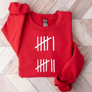 May include: A red crewneck sweatshirt with white tally marks printed on the front. The design consists of two sets of vertical lines, each with a diagonal slash. The sweatshirt is folded, with the sleeves visible.