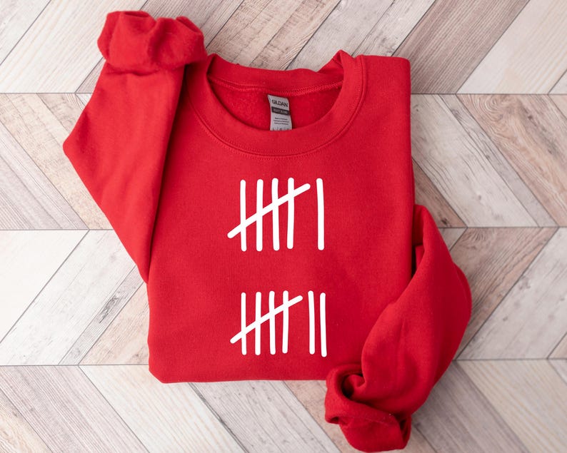 May include: A red crewneck sweatshirt with white tally marks printed on the front. The design consists of two sets of vertical lines, each with a diagonal slash. The sweatshirt is folded, with the sleeves visible.