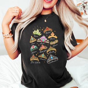 May include: Black t-shirt featuring colorful illustrations of various princess tiaras, each labeled with a name like "Belle," "Cinderella," and "Snow White." The tiaras are adorned with gems and floral designs.