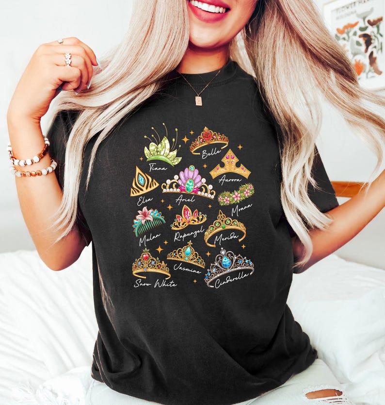 May include: Black t-shirt featuring colorful illustrations of various princess tiaras, each labeled with a name like "Belle," "Cinderella," and "Snow White." The tiaras are adorned with gems and floral designs.