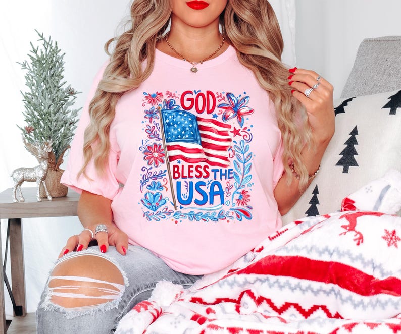 May include: A light pink short-sleeve t-shirt featuring a graphic print of an American flag with the text "GOD BLESS THE USA" in blue and red lettering, surrounded by a decorative border of red and blue floral and leaf illustrations.