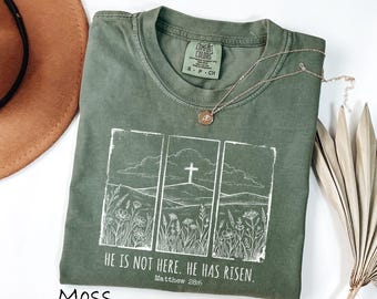 Op de afbeelding: Een mosgroen t-shirt met een witte afbeelding van een kruis op een heuvel, wilde bloemen en de tekst "HE IS NOT HERE. HE HAS RISEN. Matthew 28:6." Een gouden ketting ligt op het shirt.