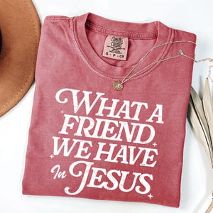 K&ouml;nnte beinhalten: Ein rotes T-Shirt mit einer wei&szlig;en Grafik im Vintage-Stil, auf dem "What a Friend We Have in Jesus" steht.
